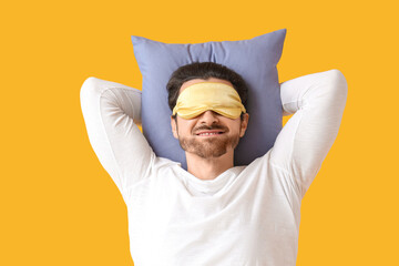 Handsome young man with sleep mask and pillow sleeping on yellow background