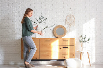 Young woman with eucalyptus branches in vases near chest of drawers in room