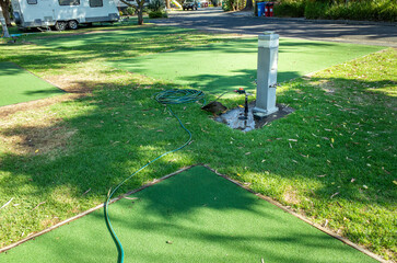 A water service point stands on an artificial green turf surface at a caravan park, with a water hose coiled nearby for camper access. Well-maintained grass areas on a camping ground with campsites.