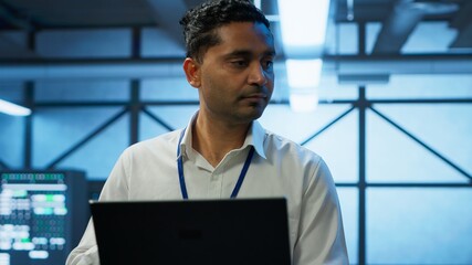 Fototapeta premium System administrator in data center using notebook to design, implement and manage gear, ensuring optimal performance and reliability. Indian man in server room coding on laptop, camera A