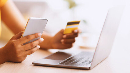 Mobile Banking App. Unrecognizable african american woman holding debit credit card and cellphone, sitting at table with laptop, cheking her online wallet. Blurred background, selective focus