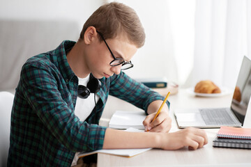 Serious schooler doing homework at his room, writing in notebook, using laptop, side view, empty...