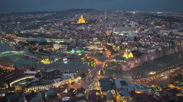 Fascinating aerial city view of Tbilisi in the evening. The illuminated Sameba Cathedral, the Kura River, the Peace Bridge and the old town area offer an impressive atmosphere.