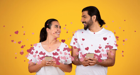 A couple stands together against a bright yellow background, smiling and exchanging affectionate messages on their smartphones. Heart graphics surround them, capturing the spirit of love.