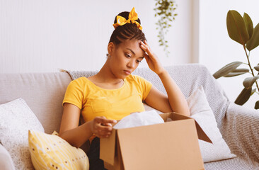 Wrong Item Concept. Sad unpset african american lady sitting on couch and unpacking cardboard box,...
