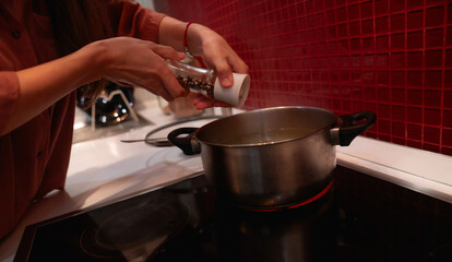 A woman is adding spices from a jar to a pot on the stove in her modern kitchen. The image captures a cozy, home cooking moment with a focus on flavor and preparation.