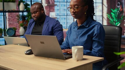 African american woman checking reports in a notebook to solve tasks with her partner, doing freelance work from the comfort of their home. Focused people doing research with laptop. Camera A.