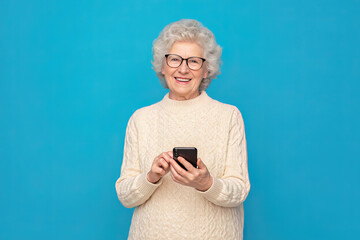 Fototapeta premium Happy aged caucasian woman grandmother holding smartphone using mobile online apps, browsing. Smiling old elderly lady texting sms message chatting with family on phone isolated over blue background..