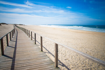 Long wooden boardwalk on sandy beach with blue sky and sea.