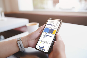 A person holding a smartphone displaying business analytics and financial data while having coffee in a café. The screen shows charts and graphs, representing data analysis.