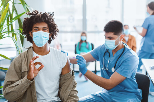 Happy African American man in face mask showing okay hand sign gesture while getting receiving vaccine in health center. Vaccination, immunization, inoculation and Coronavirus flu pandemic concept