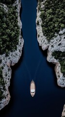 Fototapeta premium Aerial view of a boat cruising through a narrow, serene blue waterway surrounded by lush green forest-covered cliffs