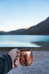 A solitary figure stands by the serene waters of El Yeso, Andes, Chile The hand, weathered and aged, clasps a copper mug, reflecting the pale light of the dawn in the mountains