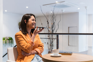Happy woman uses cellphone for communication over speaker records voice message in modern cafe.