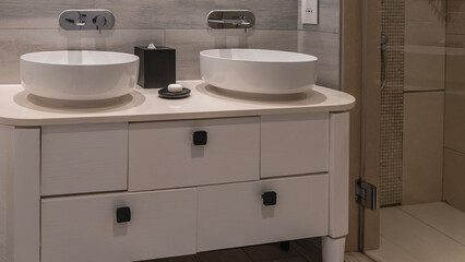 Bathroom interior details in light beige and cream tones. Two round ceramic wash basins. Commode with drawers. There is a soap dish, a box of napkins on the countertop. Metal faucets on a tiled wall.