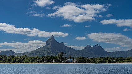 The picturesque coast of a tropical island. Mountains with sharp peaks against a blue sky, clouds. Green vegetation on the shore. Cottages, straw umbrellas, a sandy beach are visible.The ocean is calm