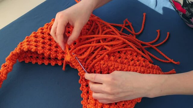 Hands carefully crafting macrame bag with orange cords. Bohemian-inspired decor coming to life, highlighting the art of handmade design and the creative DIY process. Female hobby.