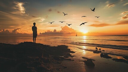 A silhouette of a person standing on a rock overlooking a beautiful sunset over the ocean, with seagulls flying in the sky.