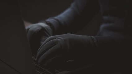 Closeup of man wearing glove typing on computer notebook in concept of criminal hacker trying to hack into victim laptop to steel confidential information in cybersecurity.