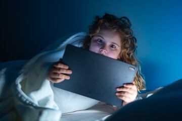 Child using Tablet in bed at night. Kid in pajamas using tablet before going to bed. Kid using gadgets. Child watching his tablet in bed. Kid lying on bed and surfing Internet on tablet in dark room.