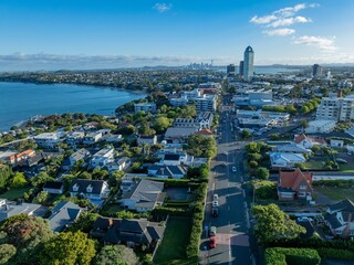 High-angle view of a vibrant city neighborhood. Residential and commercial buildings line streets, with cars parked. A beautiful day. TAKAPUNA, AUCKLAND, NZ