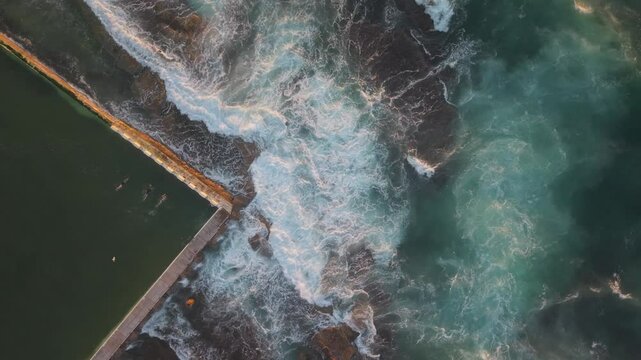 A top down aerial static of waves crashing at Merewether Ocean Baths, vibrant textures as swimmers enjoy pool