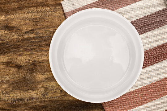 A white ceramic plate placed on a rustic wooden table with a striped cloth napkin. Top view of minimalist tableware. - Powered by Adobe