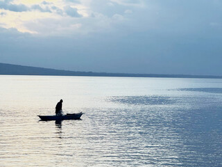 Obraz premium A peaceful scene featuring a solitary fisherman captured in silhouette against the calm lake waters under a soft fading sky.