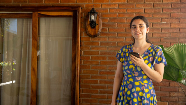 Modern Lifestyle: A portrait of a smiling woman holding a phone, she is standing against a brick wall, embodying contemporary life. The sun gently illuminates the scene.
