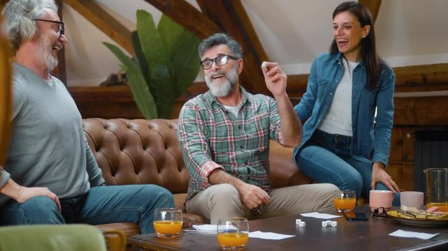 Group of retired senior friends are having fun at home playing board games, a proud elderly man is holding a dice and bragging about the point he won and the victory