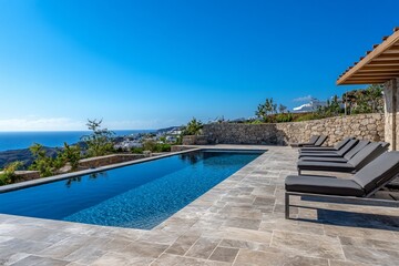 Fototapeta premium Luxury infinity pool overlooking Aegean Sea, sun loungers, stone terrace, clear sky.