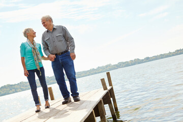 Fototapeta premium Happy, senior and couple walking on pier for retirement, vacation journey and travel destination. Below, elderly people and holding hands for marriage commitment, holiday location and bonding on lake