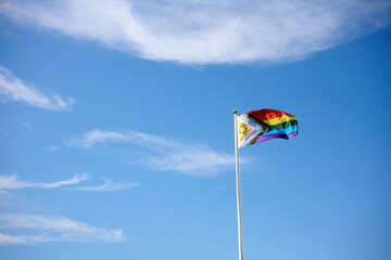 Progress pride flag waving on a flagpole against blue sky with clouds