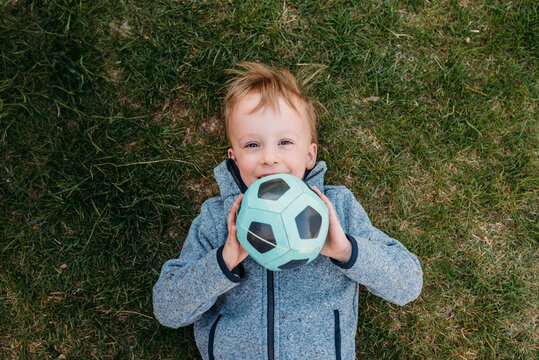 Smiling boy lying on grass holding a soccer ball playfully.