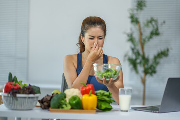 Young asian woman pinching her nose due to bad smell coming from a bowl of salad, sitting at the...