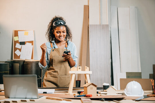 Carpenter woman in a workshop uses her smartphone. Business owner and product designer managing projects online, reviewing designs, and gathering feedback for evaluation and improvement