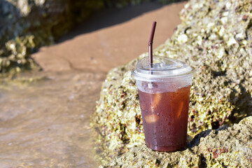 Soft Drink at beach, coffee with ice and straw in plastic glass at beach, Close up of take away plastic cup of iced black coffee (Americano) with drop, Black coffee in a plastic cup on the beach