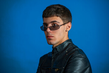 Young man wearing sunglasses and leather jacket posing on blue background