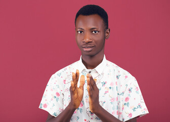 A young man in a floral shirt claps his hands together with a cheerful expression against a coral...