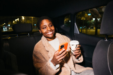 Cheerful young woman using her smartphone and drinking coffee while traveling in taxi at night