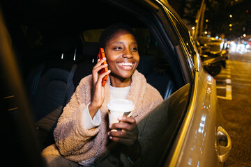 Cheerful young woman talking on mobile phone and drinking coffee while sitting in backseat of car...