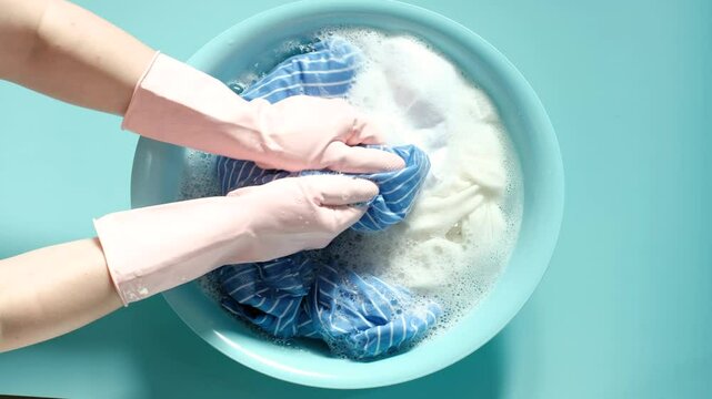 Female hands in rubber gloves washing clothes in basin on blue background, top view.