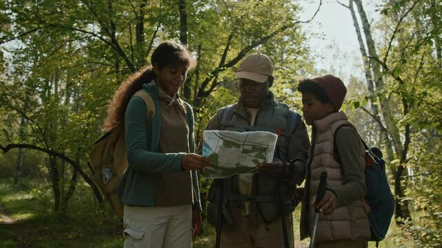 African American family of father and two teenage children wearing backpacks looking at map discussing route together while hiking in woods on sunny day