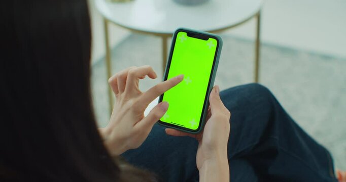 Back view of woman at smartphone with green screen for copy space. Chromakey mock up with tracking markers. Asian lady watching video news on couch close up. Tap to click on centre of screen.