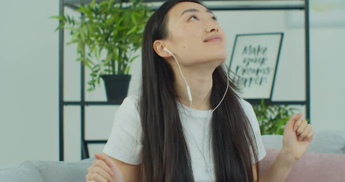 Portrait of Asian woman having fun while listening to music at home. Attractive young female in headphones dacing in living room.