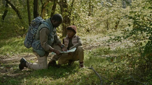 African American father down on one knee with backpack and gen alpha son reading map together when hiking on trail in early fall