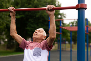 Elderly man pulls himself up on a horizontal bar on street sports field