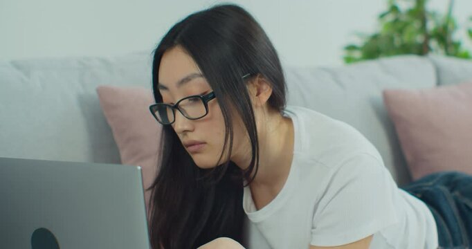 Pretty Asian woman in glasses lying on the sofa in living room using laptop computer, chatting use social media network, watch received video from friend.
