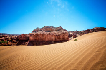 Golden sand dunes stretch endlessly, leading to towering desert cliffs beneath a vast blue sky. A breathtaking view of nature’s raw beauty, capturing the essence of Mongolia’s arid landscapes
