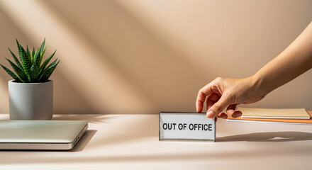 Hand placing out of office sign on desk next to laptop and plant during work hours